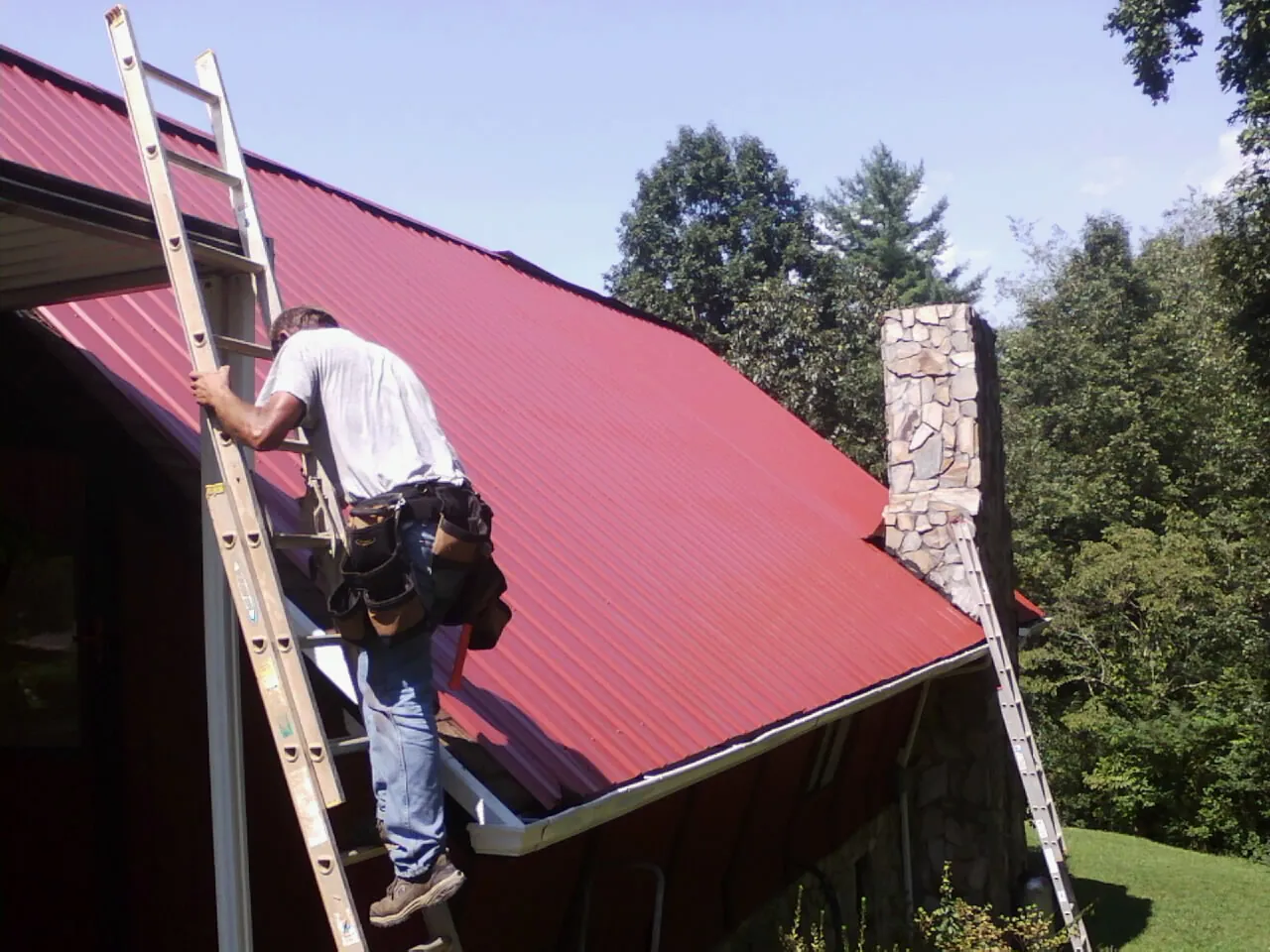 Roofer on a ladder beside a red metal roof and stone chimney — Soffit & Fascia Repair by Roofing Brothers in Hartstown